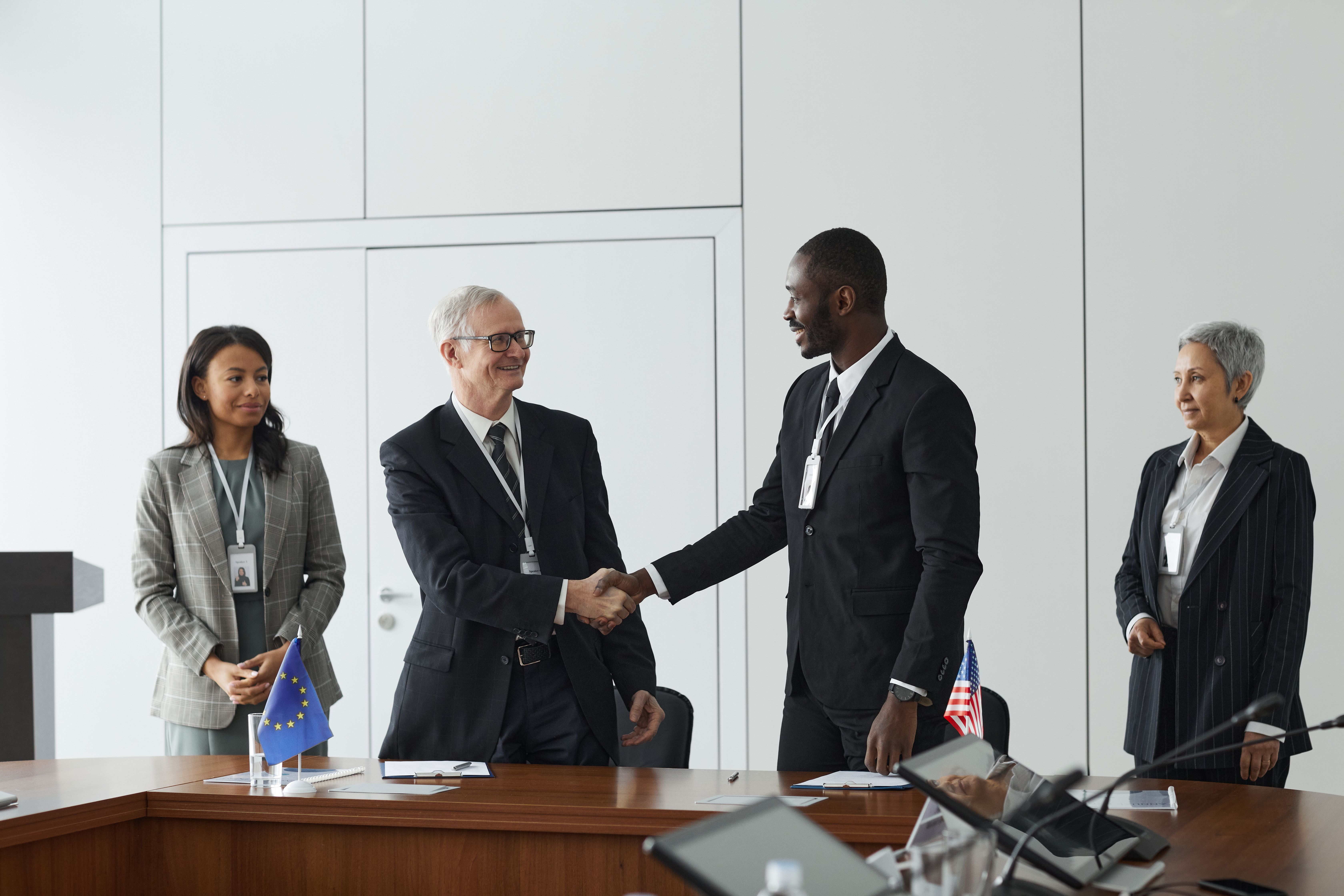Two officials shaking hands in a conference room with national flags, symbolizing international cooperation