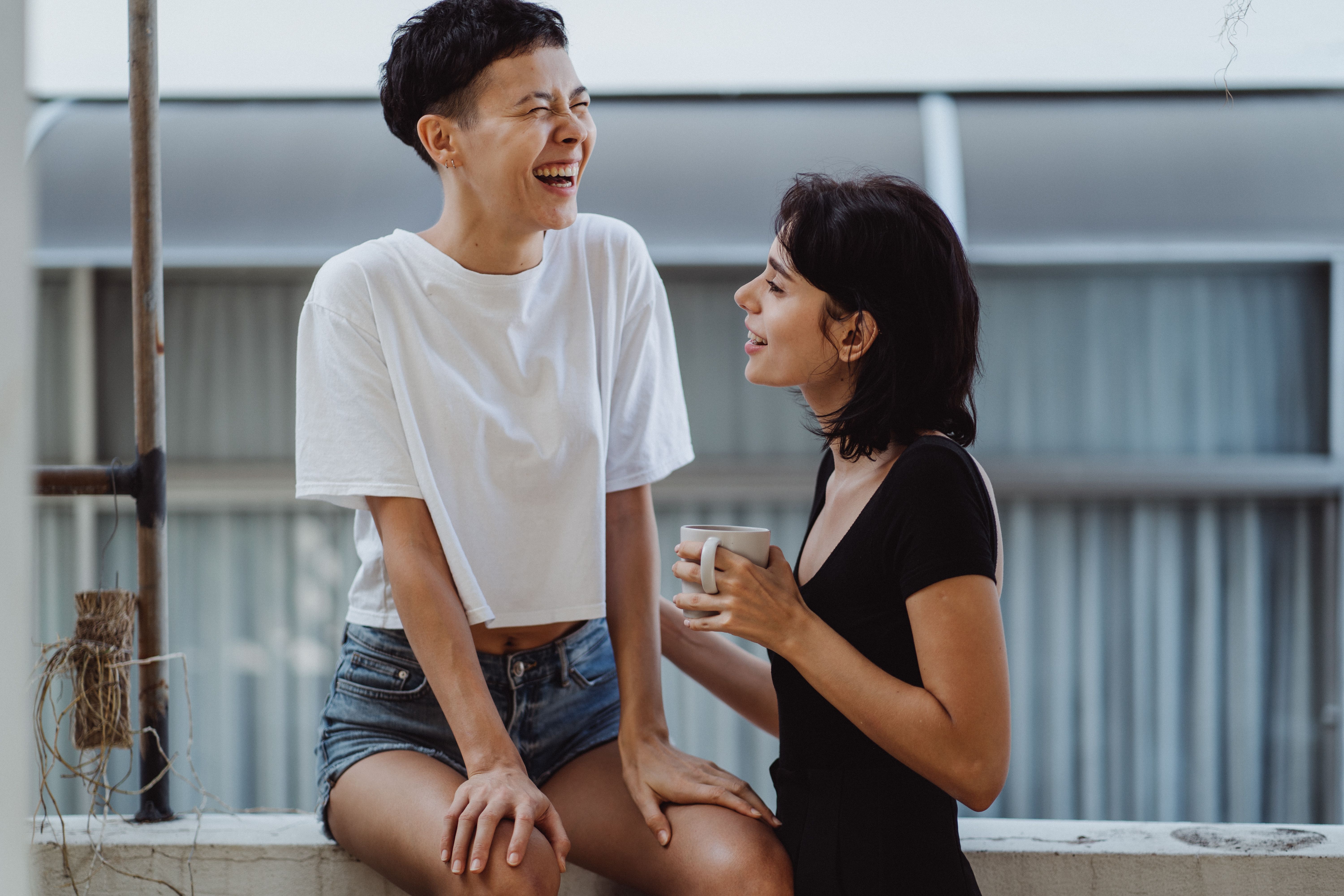 Two women laughing over coffee on a balcony, conveying friendship, warmth, and everyday connection.